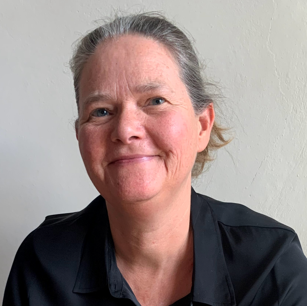 Close-up portrait of a woman with grey hair tied back and blue eyes, smiling gently against a plain off-white background.